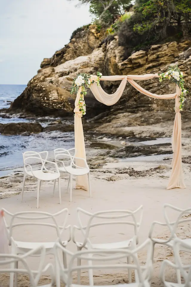 Arco de tul y flores junto a las rocas, con las sillas blancas preparadas para la ceremonia