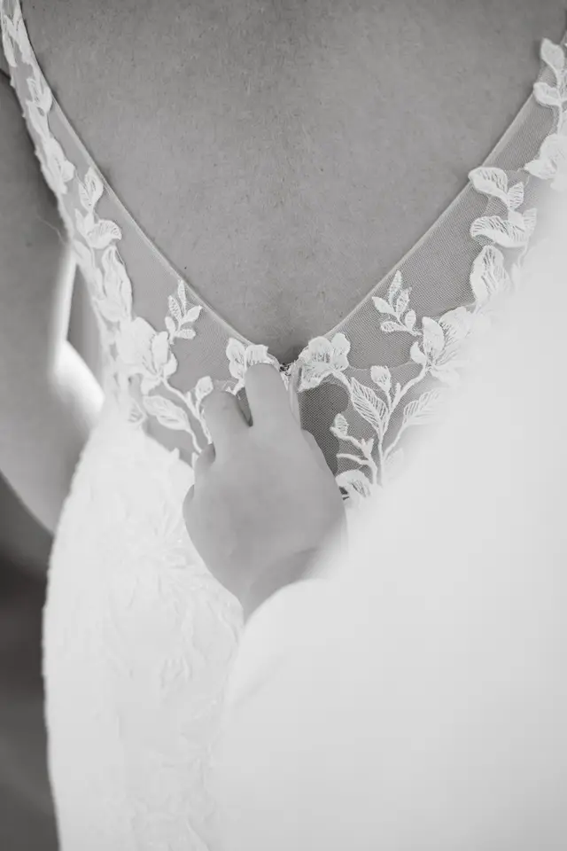 Detalle de la espalda de encaje del vestido mientras abrochan a la novia, en blanco y negro