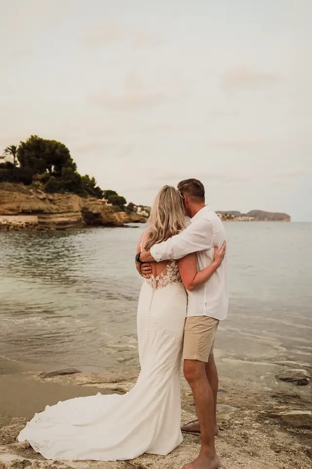 Los novios abrazados en la orilla, con la cala al fondo