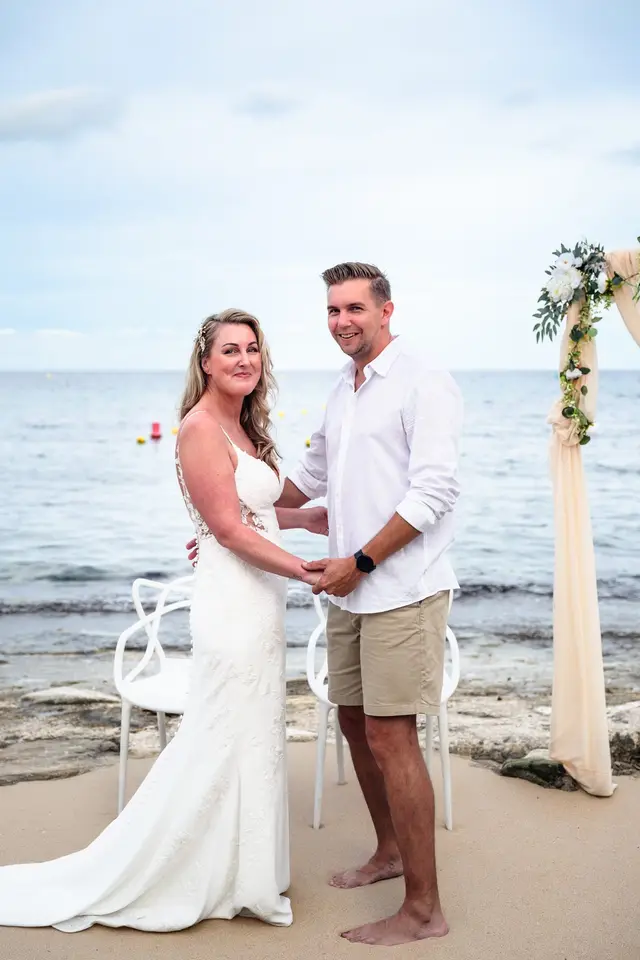 Los novios, de la mano junto al arco, sonríen frente al mar