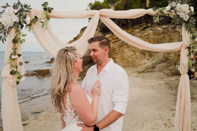 Los novios se miran sonrientes bajo el arco de tul y flores