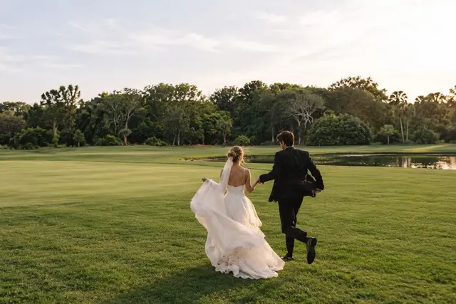 Los novios corren de la mano por el césped al atardecer, con el velo y la falda del vestido al viento